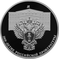 На переднем плане реверса монеты изображён герб, на фоне — здание Прокуратуры России в Москве