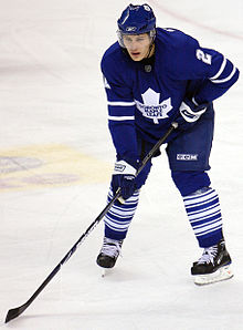 A full body shot of a young man wearing a blue jersey, blue hockey pants and skates.