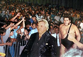 Bobby Heenan (front), a Caucasian man in a black sequin jacket, leads André the Giant to the ring