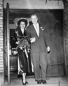 A man in a suit leads a flower-carrying woman by the hand, walking out of a chapel.