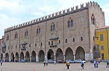 A long, grey stone building with a line of arches at ground level, above which is a row of small windows interspersed with balconies. Above this row is a line of larger windows below a crenellated roof line.