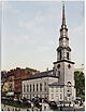 Ground-level view of a brick church with a large, white, tapering spire; a brown skyscraper is visible in the distance, with several shorter high-rises located closer to the church.