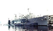 Three submarines are moored side-by-side to a large surface ship, which in turn is held to a dock in the background.