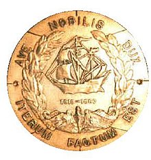 A round metal object features a sailing ship in the center. In a circle around the edges are the Latin words «Ave Nobilis Dux, Iterum Factum Est».