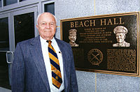 Elderly man in coat and tie standing next to a bronze dedication plaque located at the building entrance.