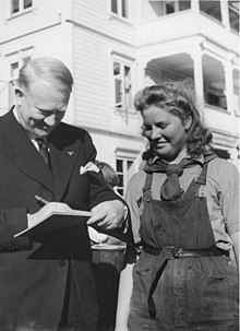 A middle-aged man in a dark coloured suit is writing on a pad of paper. Standing close to him and watching is a woman in her late twenties to earlier thirties. She is smiling, and wearing dark-coloured dungarees, with a shirt underneath and a scarf tied around her neck.