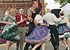 A group of dancers in colorful Western clothing promenading in a circle, with a man speaking into a microphone on a stage in the background.