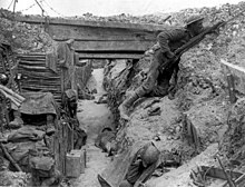 Soldier on guard armed with a rifle wearing a Brodie helmet, three other men are asleep in the bottom of the trench