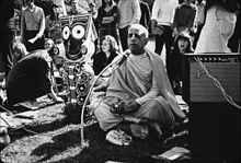 A black-and-white photo of the swami sitting cross-legged with a figure of round-eyed smiling deity to his right