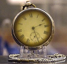 Фотография латунных карманных часов на подставке с серебряной цепочкой, обвивающей основание. Стрелки часов показывают 2:28.