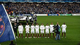 group of several men in several rows, standing, dressed in black and white football kit, flanked by man with camera