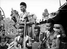 Black and white photo of five shirtless men of Melanesian ethnicity wearing military webbing. Three of the men are armed with guns, with one also carrying a large knife.