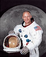 A portrait of a man wearing an space suit, with his helmet on the table in front of him. Behind him there is a large photograph of the lunar surface.