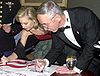 A young woman and an older man, both dressed in formal wear, leaning over to sign a poster.