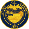 A navy blue seal with gold lettering and imagery. The seal contains a shield, supported by 33 stars and with an eagle with its wings spread on top; "STATE OF OREGON" is written above the eagle and "1859" appears below the shield. Within the shield appear a sun with its rays extending to two ships and a mountain and trees, two oxen pulling a covered wagon, and a ribbon containing "THE UNION".