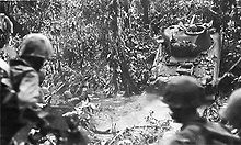 In the foreground Marines watch as a light tank climbs up the bank of creek in a jungle setting.