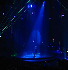 Faraway image of a female performer. She is walking through a water screen that is falling from the ceiling. A blue spotlight is on her. She is wearing blue jeans and a plastic cowboy hat. Green laser lights surround the stage.