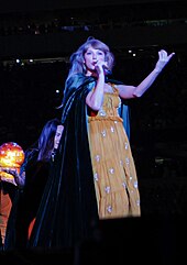 Taylor Swift performing, dressed in a black hood and orange dress
