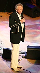 A grey-haired man in a black jacket and white pants, singing into a microphone