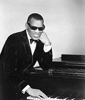 A dark-skinned man wearing dark glasses and a tuxedo, leaning on a piano