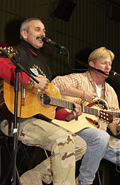 A grey-haired man with a moustache playing the guitar and singing into a microphone. A blond-haired man, also playing a guitar, is in the background.