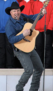 A man in a black cowboy hat, blue shirt and jeans, playing a guitar and singing into a microphone