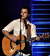 A young man with dark hair wearing a white shirt, playing a guitar and singing into a microphone