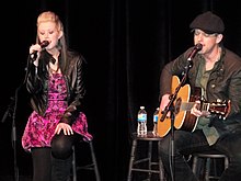 A woman with long blonde hair, wearing a purple dress, singing into a microphone, and a man wearing a cap singing and playing a guitar