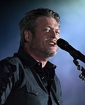 A man with greying hair and beard stubble, wearing a dark shirt, singing into a microphone