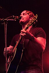 A young man wearing a dark T-shirt and cap, playing a guitar and singing into a microphone