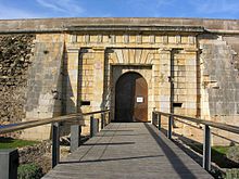 Photo shows the arched gate of a stone fortress and the bridge that approaches it.