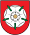 A coat of arms depicting a white flower in the middle that has a yellow stamen and green leaves all on a red background