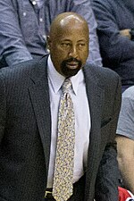 A man, wearing a brown suit and white shirt, is shouting while standing on a basketball court, surrounded by several other people.