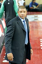 A man, wearing white shirt with a logo and the word "NBA PLAYERS" on the front, is standing and posing for a photo.