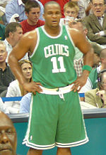 A basketball player stands on a basketball court. He wears a green jersey with the word «CELTICS» and the number 11 on the front.