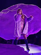 Taylor Swift singing onstage, dressed in a purple sparkling dress and a faux-fur coat