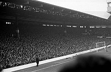 Photograph of a stand full of people. In front of the stand is a field and a goal.