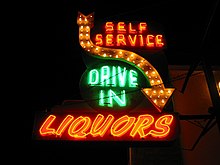 Зелёно-оранжевый неоновый знак, сообщающий о «self service drive in liquors», и имеющий направленную вниз стрелу.